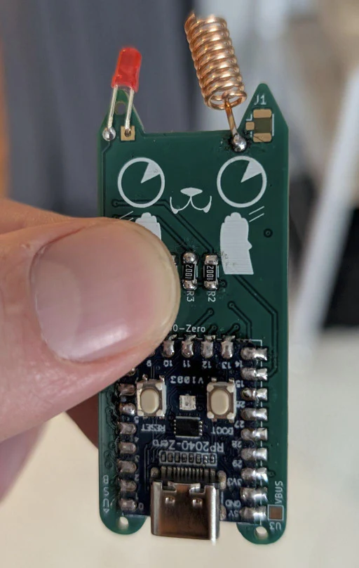 A close-up of a small electronic circuit board featuring an RP2040 microcontroller, with a copper antenna coil, an LED, and a USB-C connector. The board includes labeled pins and components like resistors and capacitors."